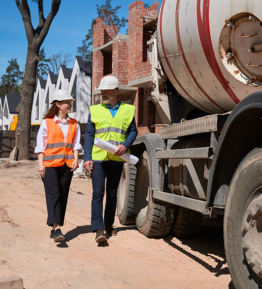 Two construction workers with helmets and vests walk near a concrete mixer at a construction site.