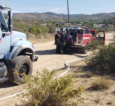 Firefighters by a red firetruck connected to a water truck on a dirt road with hills in the background.