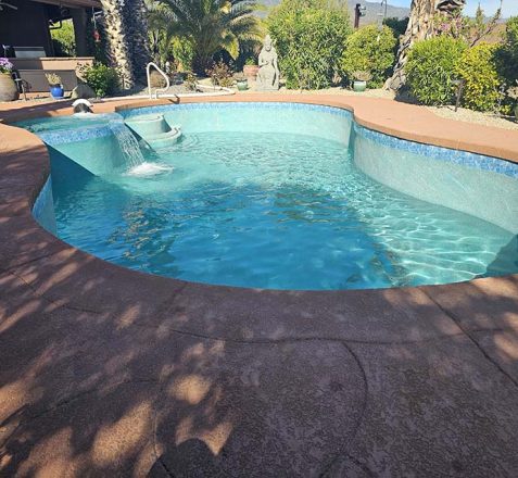 Outdoor pool with blue tiles and a fountain on a sunny day, surrounded by plants and statues.