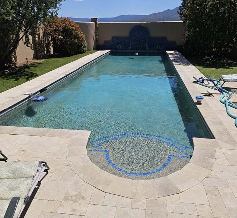 A backyard swimming pool with a blue mosaic and surrounding patio area on a sunny day.