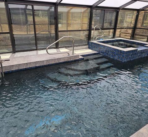 Indoor pool with a hot tub, surrounded by glass walls and a retractable roof.